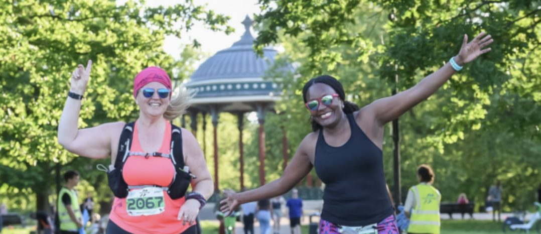 Two women are running in a park, smiling and raising their hands joyfully. Both are wearing running gear, including sunglasses, race bibs, and hydration packs. In the background, there is a gazebo surrounded by trees on a sunny day.