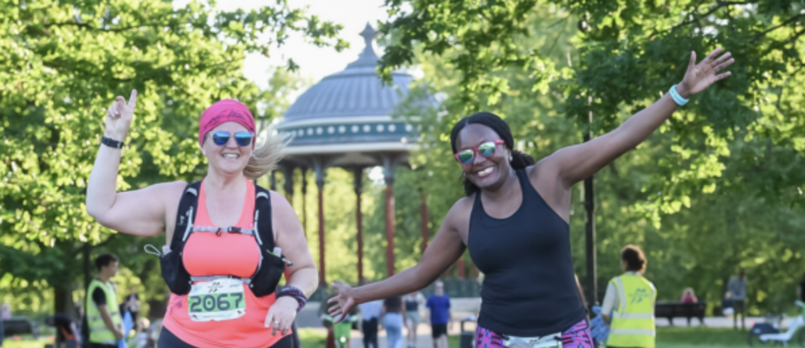 Two women are running in a park, smiling and raising their hands joyfully. Both are wearing running gear, including sunglasses, race bibs, and hydration packs. In the background, there is a gazebo surrounded by trees on a sunny day.
