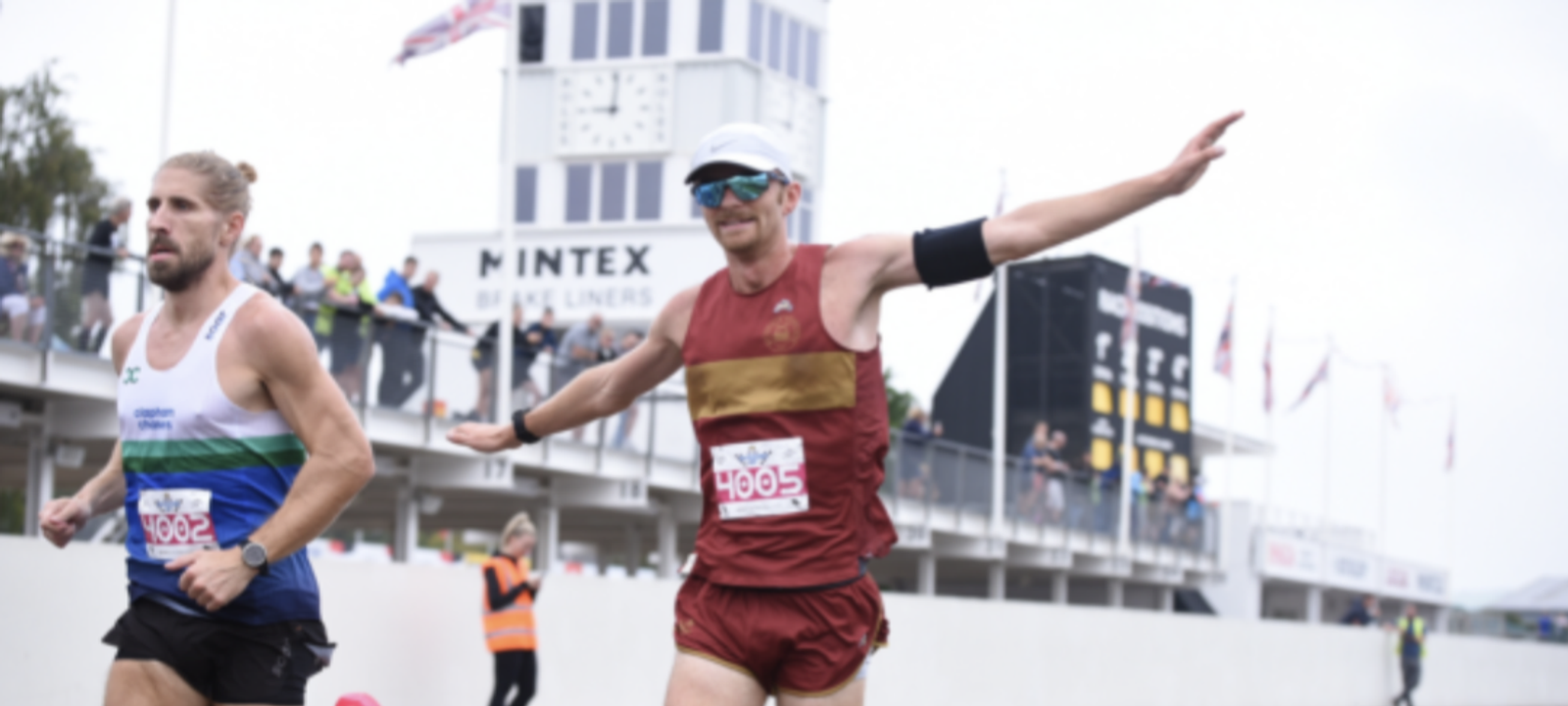 Two runners in a race approach a finish line. The runner on the left wears a white and blue tank top with black shorts, while the runner on the right wears a red outfit with yellow horizontal stripes, sunglasses, and a white cap, extending his arms out, smiling. A tower and spectators are visible in the background.