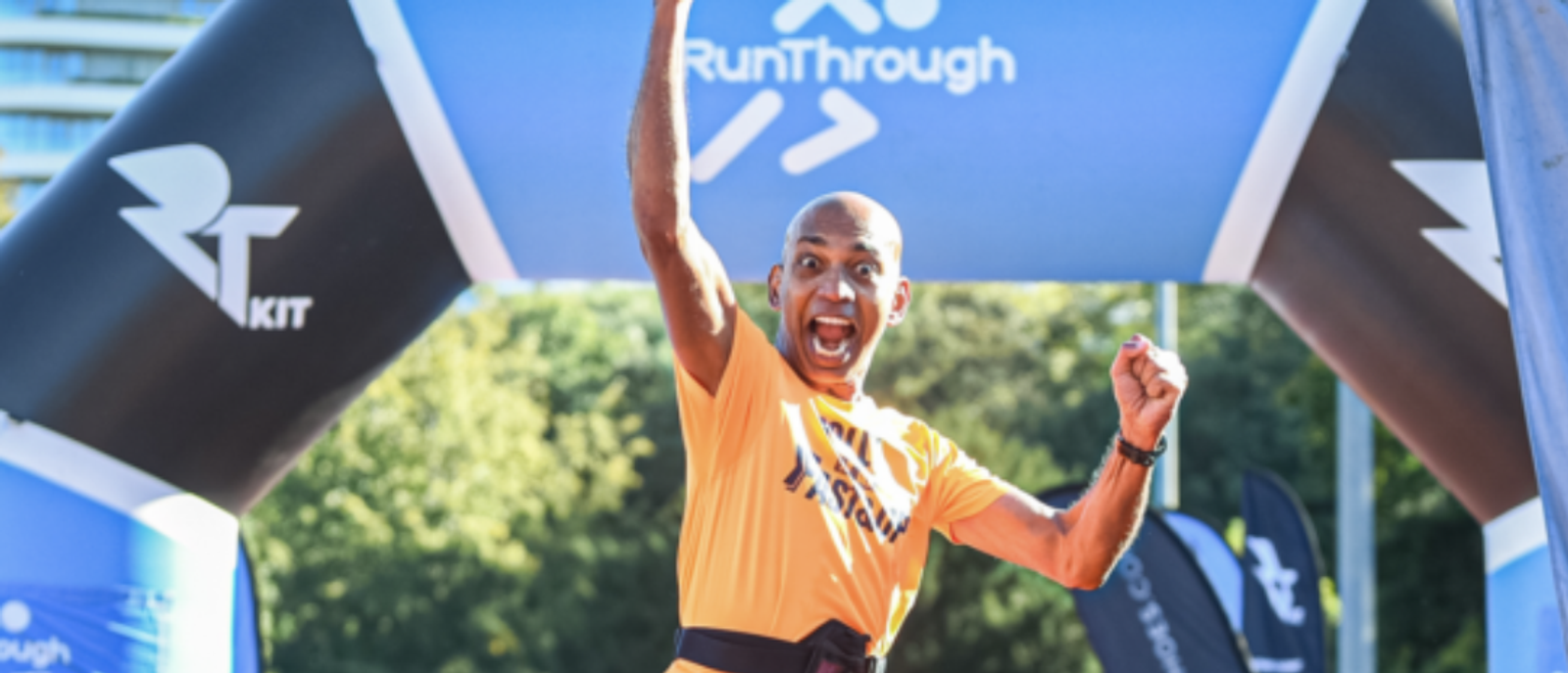 A runner in an orange shirt jubilantly raises his arm in celebration as he crosses the finish line of a race. Behind him is an archway with "RunThrough" written on it. Trees and other race banners are visible in the background.