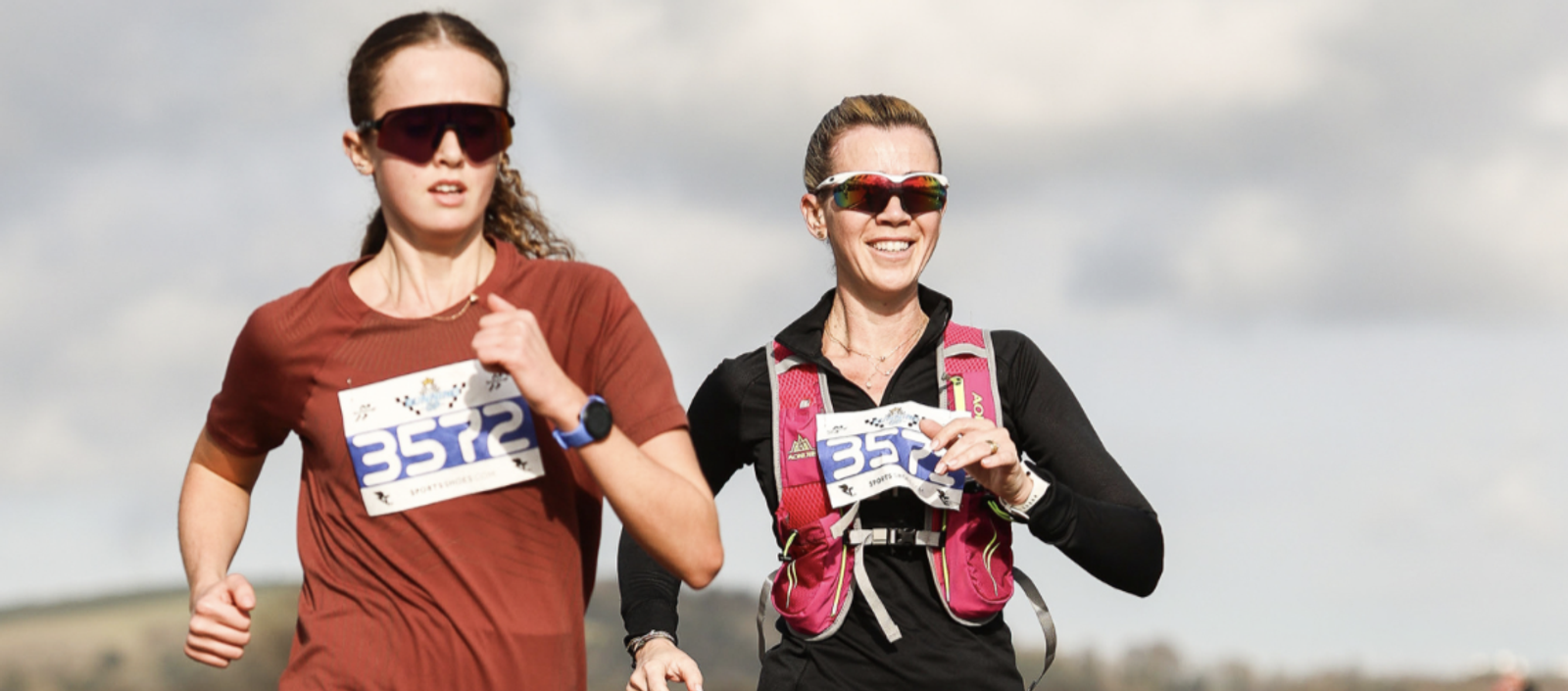 Two runners participate in an outdoor race. Both wear numbered bibs (3572) and sport sunglasses. The runner on the left wears a red shirt, while the runner on the right is in a black shirt and pink hydration vest. The sky is partly cloudy in the background.