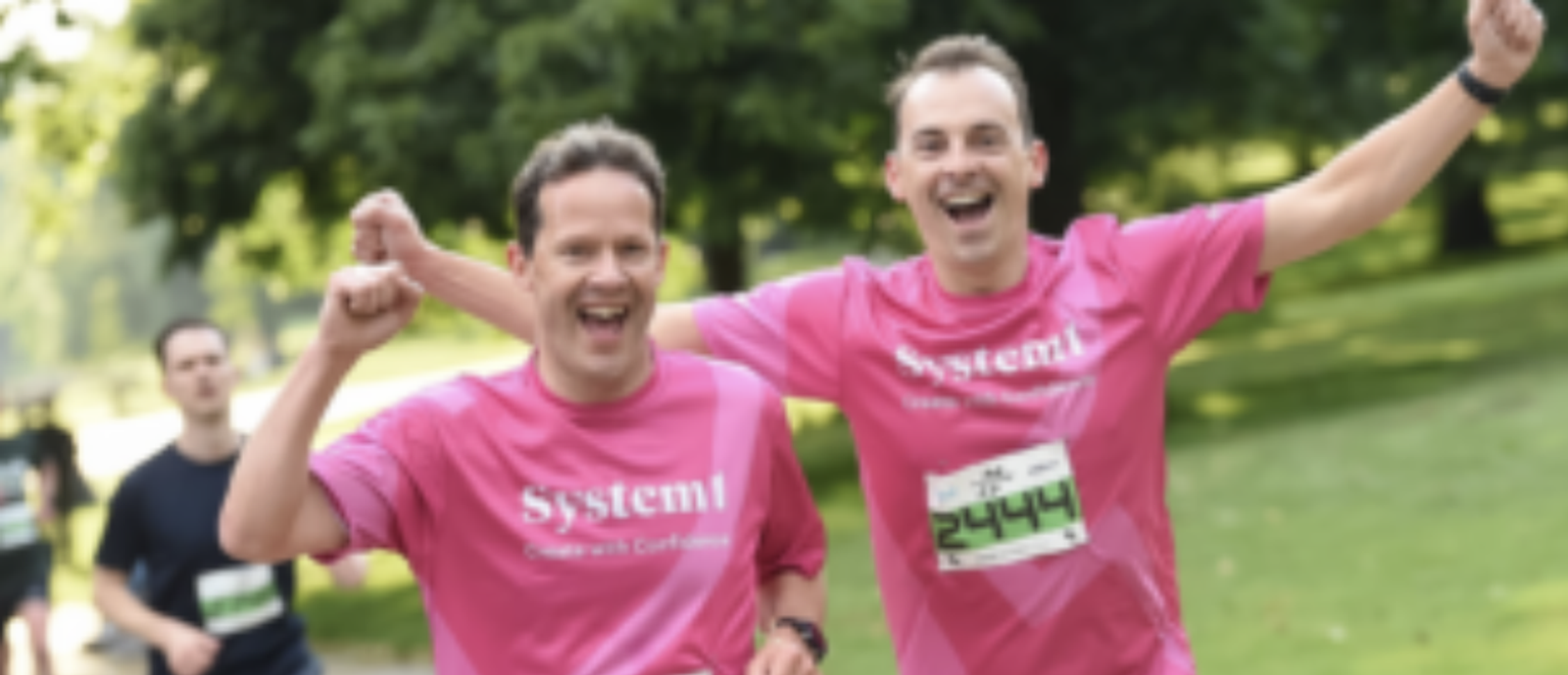 Two joyful runners in pink shirts raise their arms in celebration while running outdoors. One has a race bib numbered 2444. Green, leafy trees and other runners are visible in the blurry background.