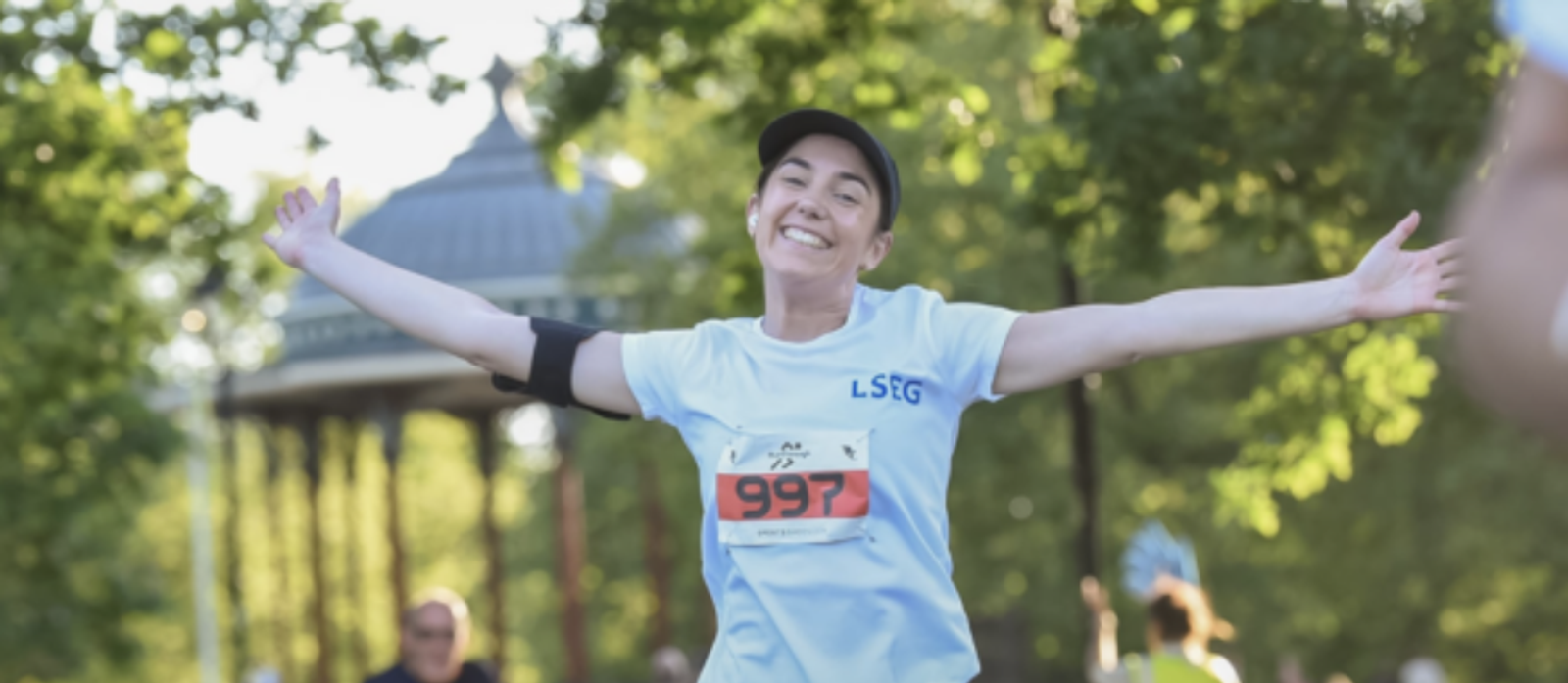 A person wearing a light blue shirt and black cap is smiling with arms outstretched while running in a park. The person has a race bib numbered 997. Trees and a gazebo are visible in the background.