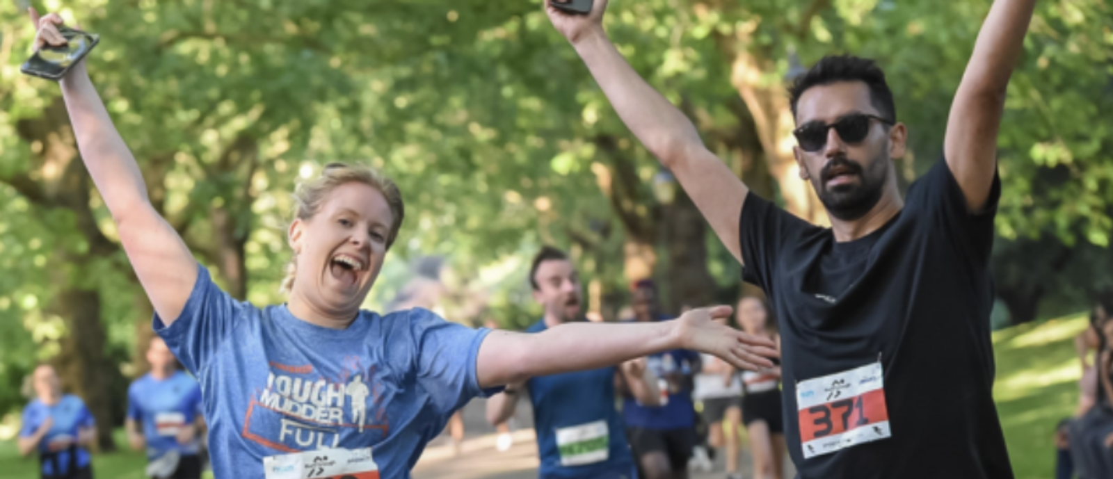 Two runners, a woman in a blue shirt and a man in a black shirt, cheerfully raise their arms while running in a park. They both have race bibs on their shirts and are smiling. Other runners can be seen in the background amidst lush green trees.