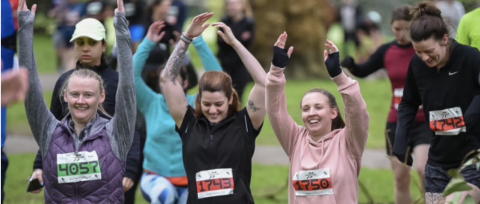 A group of runners, including three women in the foreground with numbered bibs, cheerfully raising their arms. They're participating in an outdoor event with more runners visible in the background, in a park-like setting. The mood is energetic and joyful.