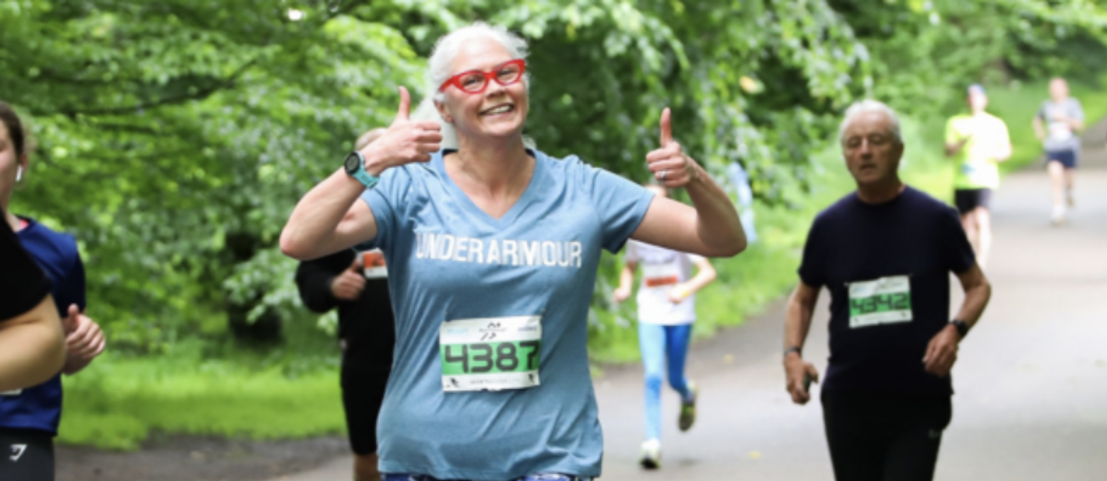 An older woman with white hair and red glasses, wearing a grey Under Armour T-shirt and a race bib numbered 4387, smiles and gives two thumbs up while running in a park. Other runners are visible in the background. The atmosphere is cheerful and green.