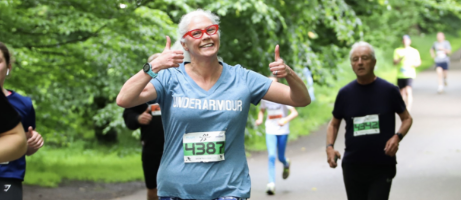 An older woman with white hair and red glasses, wearing a grey Under Armour T-shirt and a race bib numbered 4387, smiles and gives two thumbs up while running in a park. Other runners are visible in the background. The atmosphere is cheerful and green.