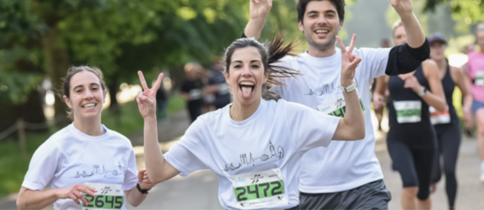 Three runners, two women and one man, are participating in a race outdoors wearing race bibs and white shirts. The woman in front is sticking her tongue out and making peace signs with both hands while smiling. The other two runners are also smiling. Trees are in the background.