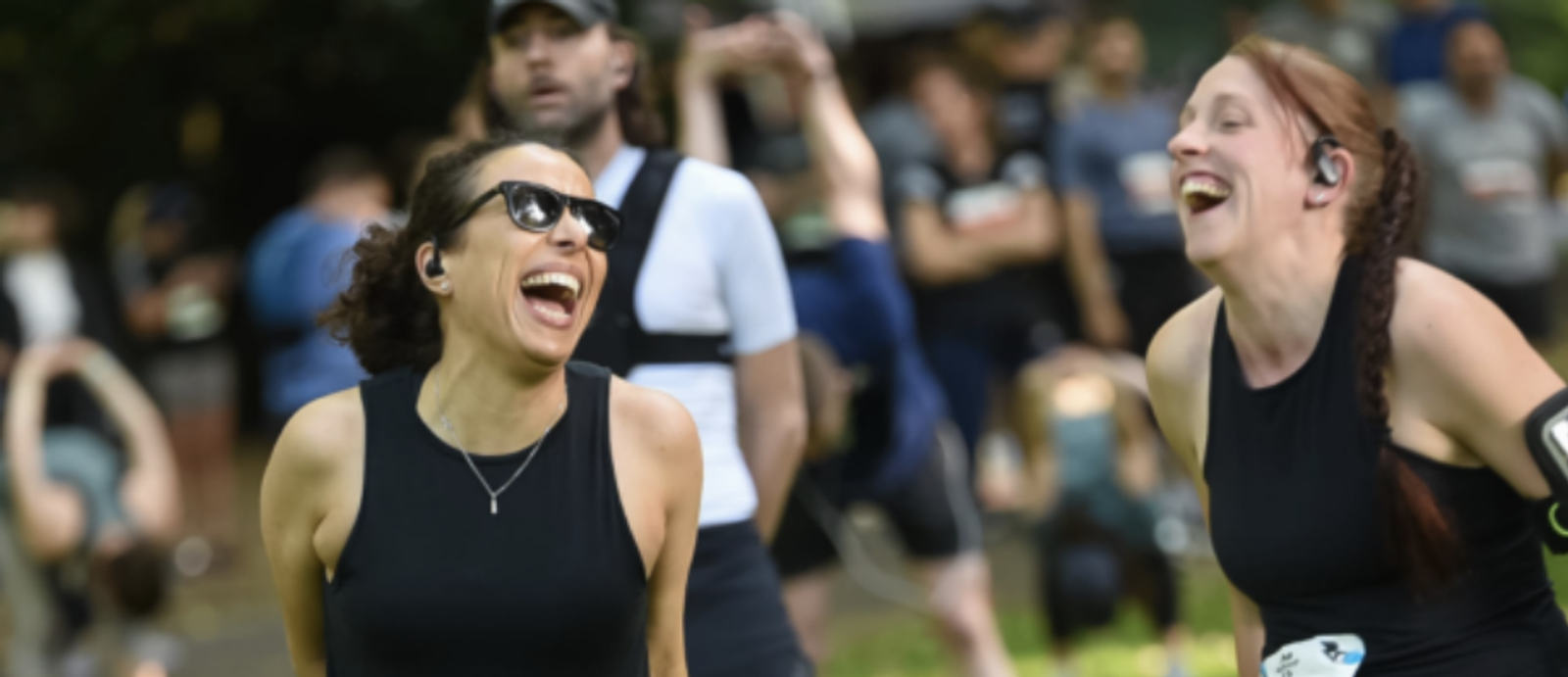 Two women are laughing together at an outdoor event. Both are wearing black athletic clothing and have earphones in. The woman on the left has sunglasses, and the woman on the right has a race bib on her waist. Other participants are in the blurred background.