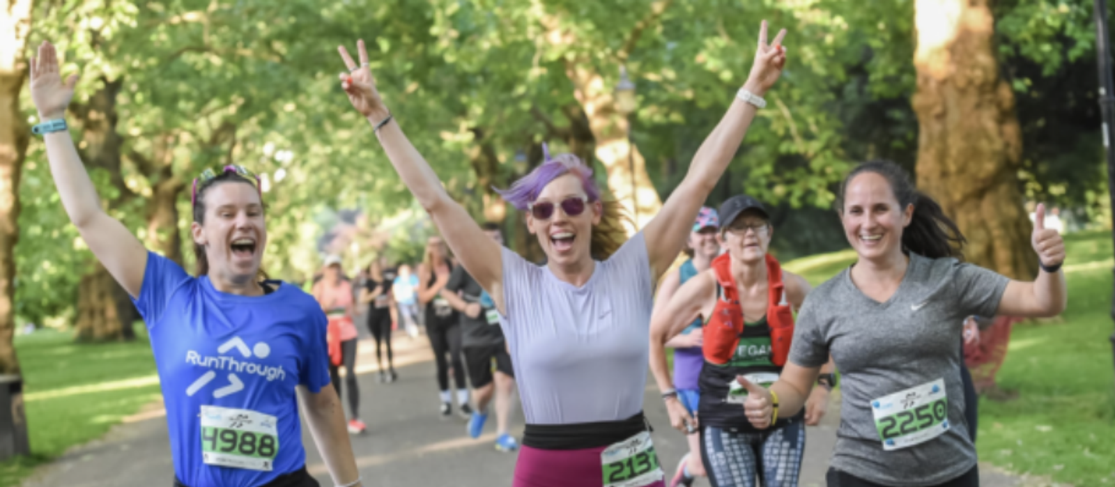 A group of enthusiastic runners smiling and making celebratory gestures while participating in a race through a park. Two women in the front hold up peace signs with outstretched arms, and other runners can be seen behind them with numbered bibs.