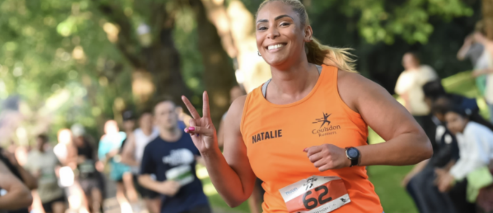 A person in an orange tank top with "Natalie" and "Guided Runners" printed on it participates in a race, holding up a peace sign and smiling. They are wearing a race bib numbered 62. The background is blurred with trees and other runners.