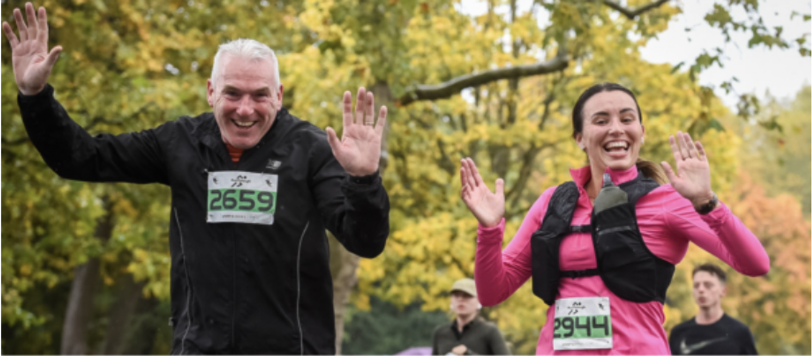 Two smiling runners wave at the camera during an outdoor race. The man on the left wears a black outfit with a bib number 2659, and the woman on the right wears a pink and black outfit with a bib number 2944. Trees with autumn leaves are in the background.