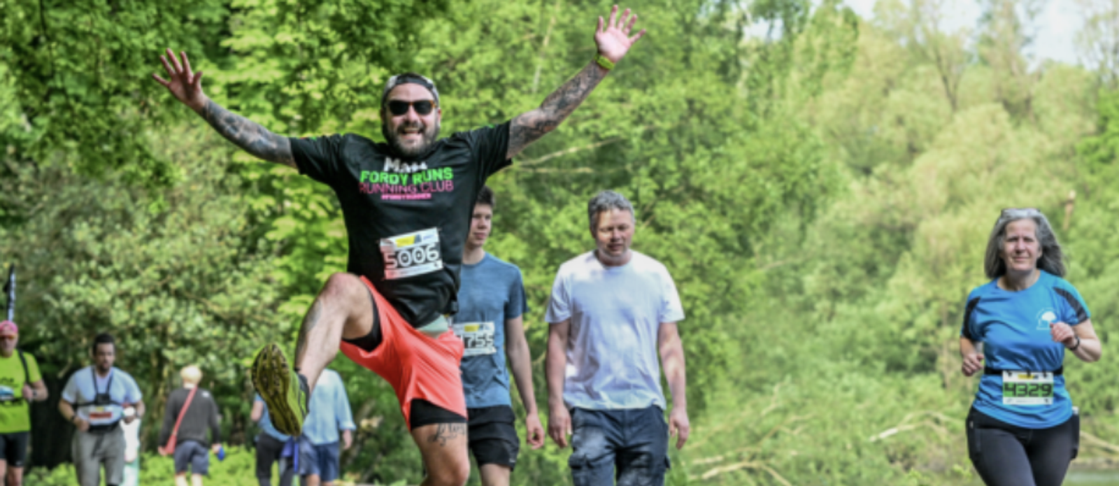 A bearded man with sunglasses, wearing a black shirt and red shorts, jumps joyfully during a forest marathon. Other participants, including a man in a white shirt and a woman in a blue shirt, walk and run along the wooded trail. Trees and foliage are in the background.
