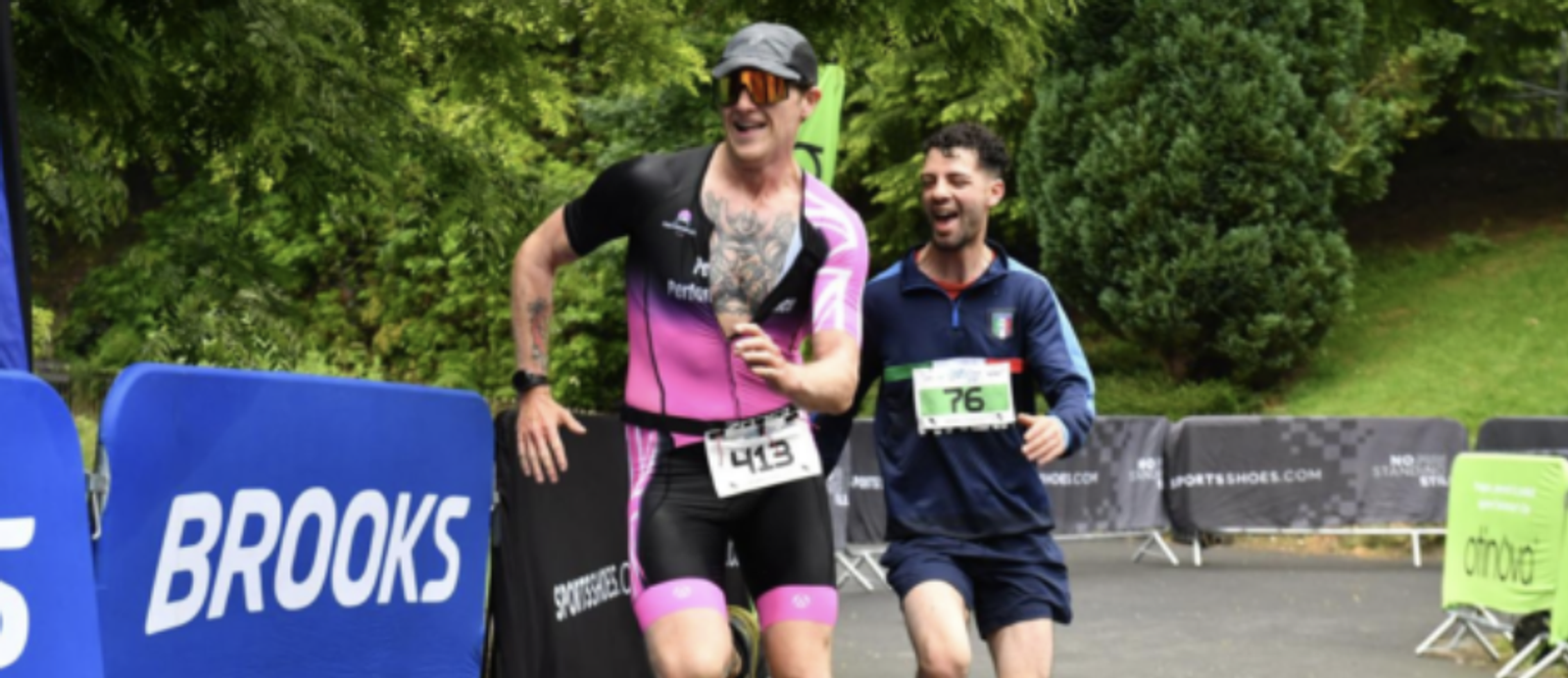 Two men are running in a race, smiling and appearing energized. The man in front wears a black and pink athletic suit with sunglasses and a cap, while the man behind follows with a blue outfit. The background shows green trees and banners, with one reading "BROOKS.