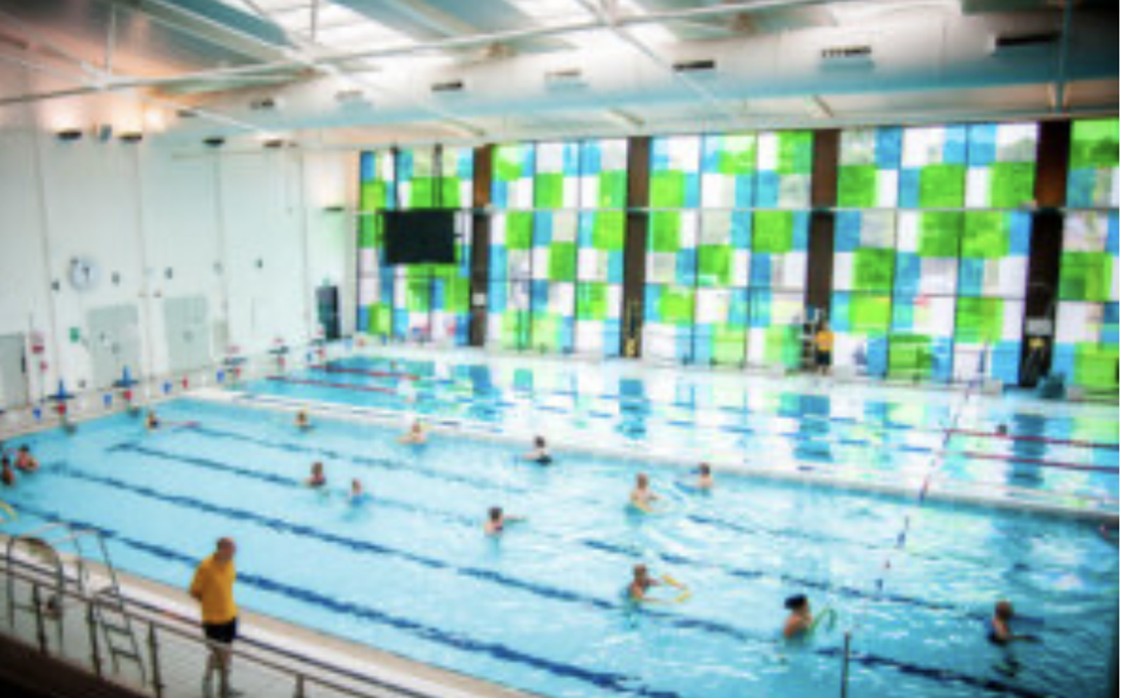 A bright indoor swimming pool with several clear lanes. People are swimming or standing in the water, engaging in various activities. A large, colorful mosaic window pane decorates one side of the pool area. The ceiling has numerous lights and ventilation systems.