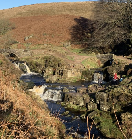 A small waterfall cascades over rocky terrain into a stream, surrounded by green and brown vegetation under a clear blue sky. Two people are seen by the water's edge, one standing and the other sitting on a rock. The backdrop consists of rolling hills and sparse trees.