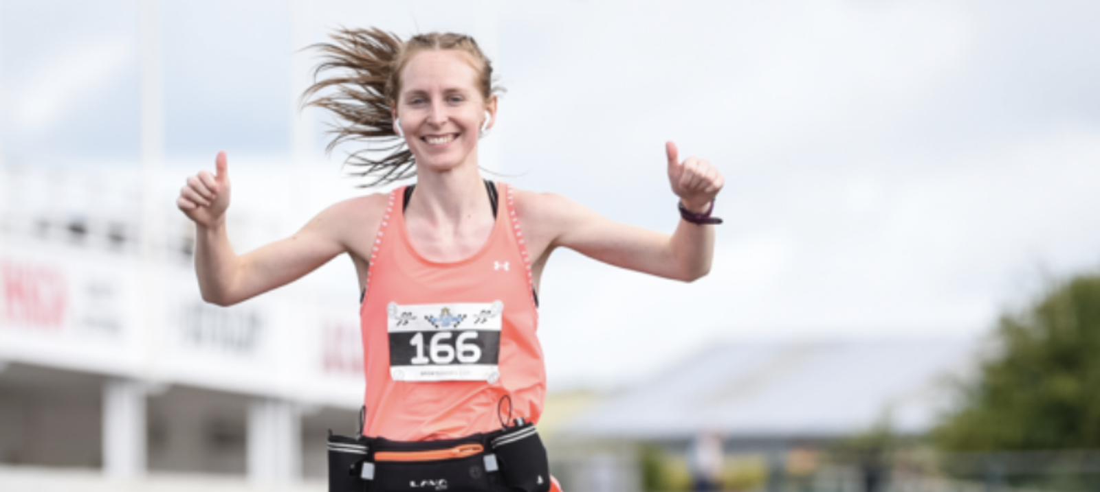 A woman with a race bib numbered 166 is smiling and giving two thumbs up while running. She is wearing a coral tank top and has a race belt around her waist. The background features a blurred outdoor setting, indicating she is participating in a race event.