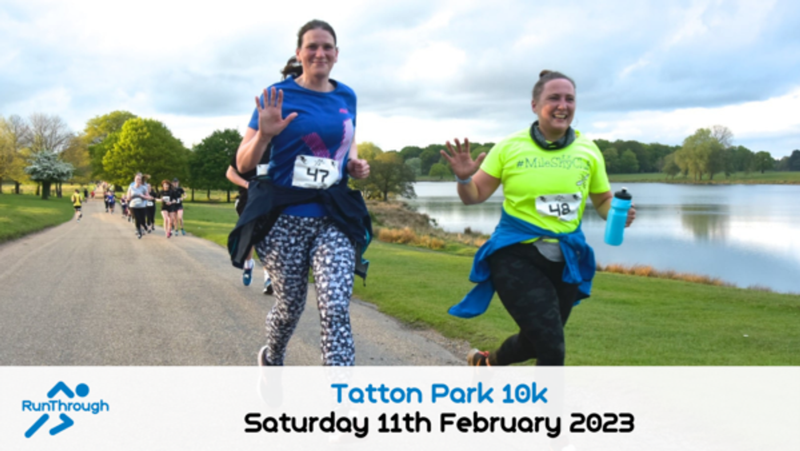 Two women smile and wave while running in a race beside a lake in a park. They wear numbered bibs and athletic clothes. Other runners follow behind on the tree-lined path. Event text and date appear at the bottom.