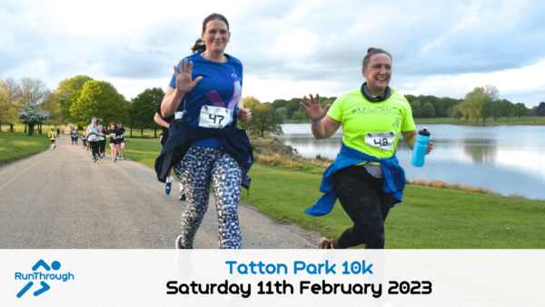 Two women smile and wave while running in a race beside a lake in a park. They wear numbered bibs and athletic clothes. Other runners follow behind on the tree-lined path. Event text and date appear at the bottom.