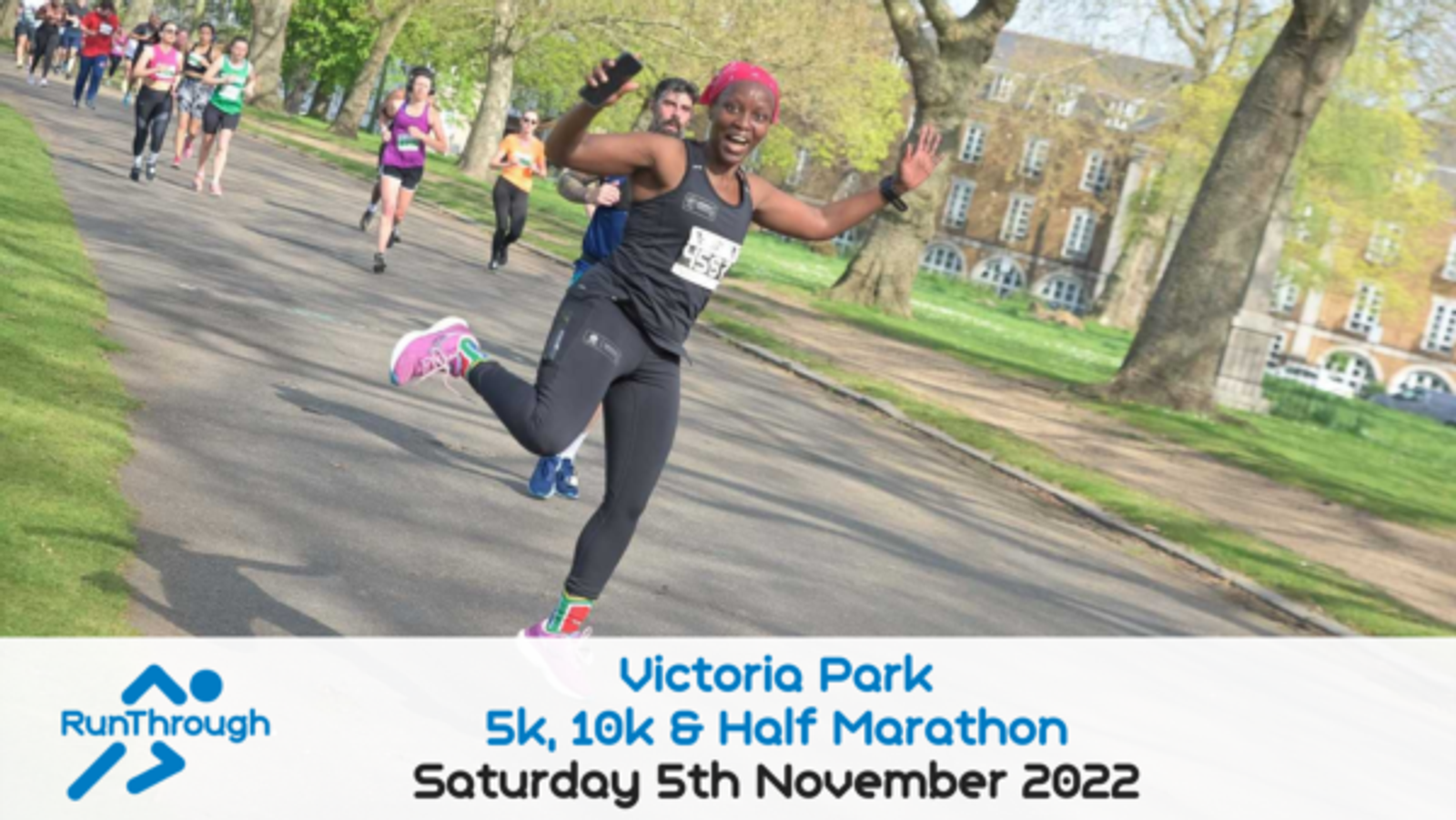 Participants running in Victoria Park during a 5k, 10k & Half Marathon event on Saturday, 5th November 2022. The foreground shows a joyful runner leaping with arms outstretched, wearing a pink headband, black top, and pants. Other runners are visible behind.