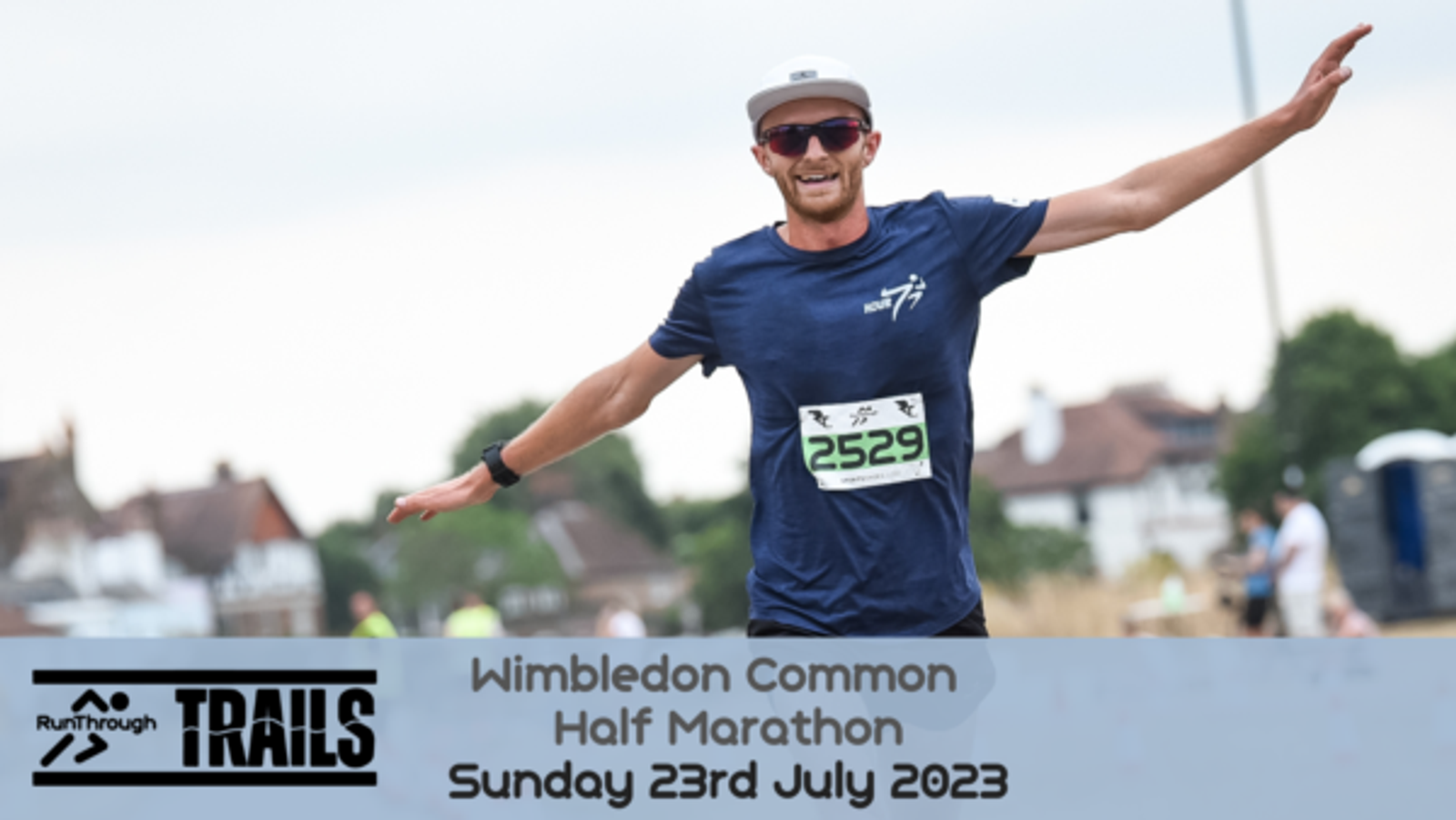 A male runner wearing sunglasses and a blue shirt with the number 2529 displayed on his chest runs with his arms outstretched. A banner at the bottom reads “Wimbledon Common Half Marathon, Sunday 23rd July 2023” with a "RunThrough Trails" logo.