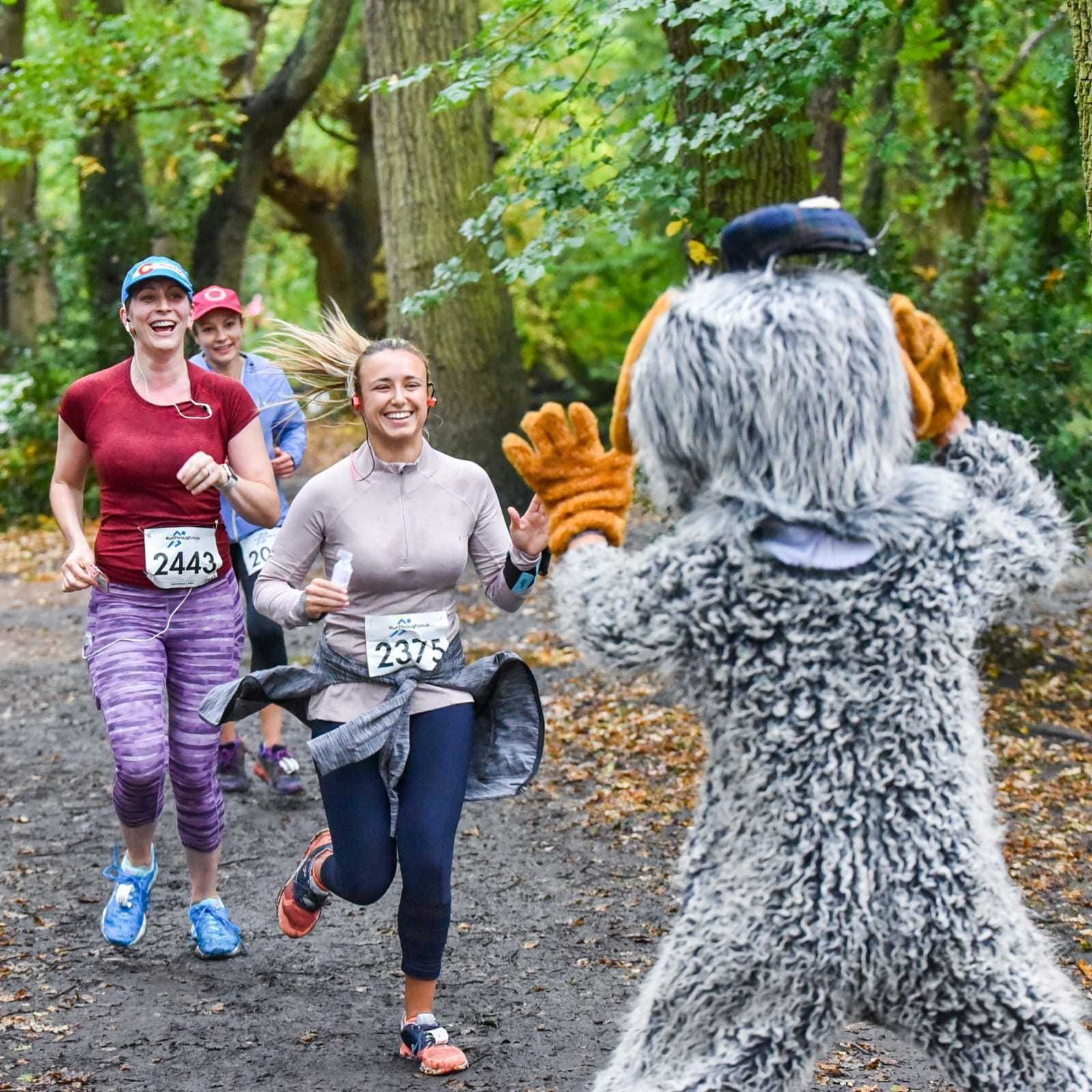 Two women wearing race bibs #2443 and #2375 run through a forested path, smiling and appearing excited. A person dressed in a furry mascot costume faces and greets them with open arms. The atmosphere is lively and filled with energy.