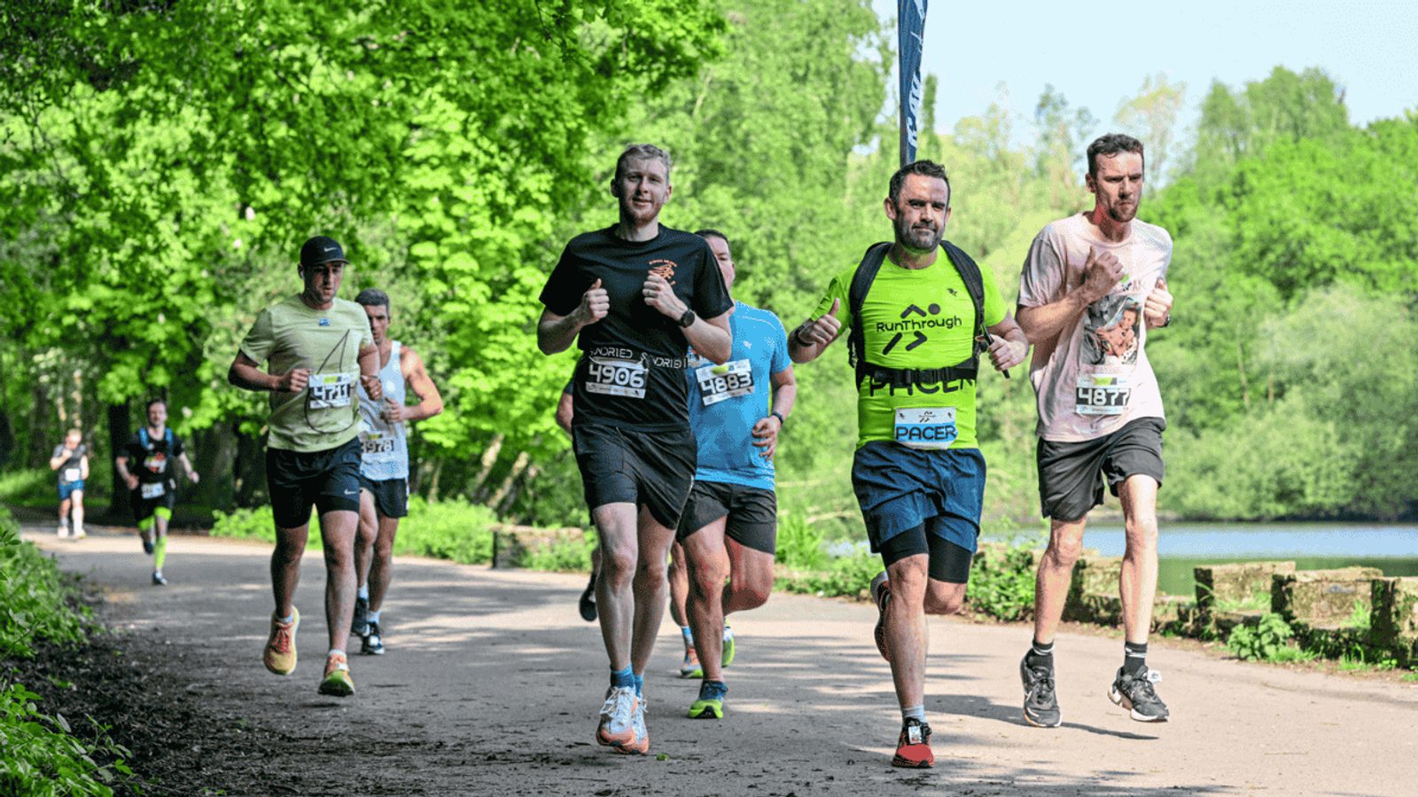A group of runners, some smiling, jog along a sunlit park path lined with trees and greenery during a race. The atmosphere is energetic and relaxed, with a lake visible in the background.