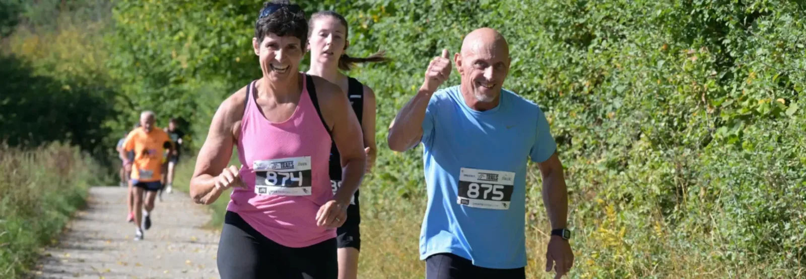 Four runners with race bibs are jogging on an outdoor trail lined with green bushes. The two front runners, a woman in a pink tank top and a man in a blue shirt, are smiling, with the man giving a thumbs-up.