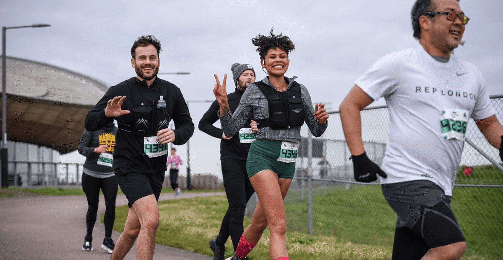 A group of runners, wearing race bibs, jogs outdoors along a paved path. Two runners smile and wave at the camera while others follow on a cloudy day near a fenced area.