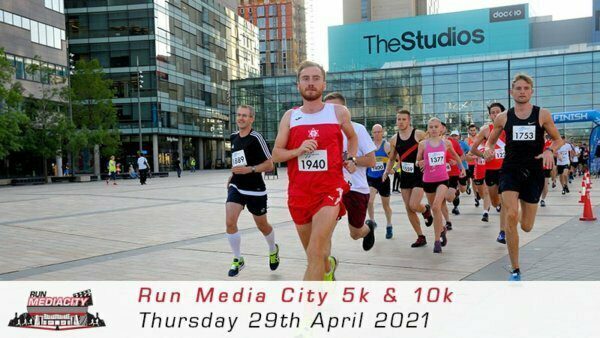 A group of runners participating in a race pass through an urban area with modern buildings and a large sign reading "The Studios" in the background. One runner in a red outfit is leading. A banner at the bottom states "Run Media City 5k & 10k Thursday 29th April 2021".