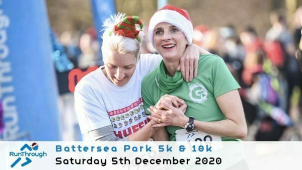 Two runners celebrate at the Battersea Park 5k & 10k event on December 5th, 2020. One wears a Santa hat and a green shirt, smiling and holding hands with another, who sports a Christmas elf hat and a white festive shirt. The event is organized by RunThrough.