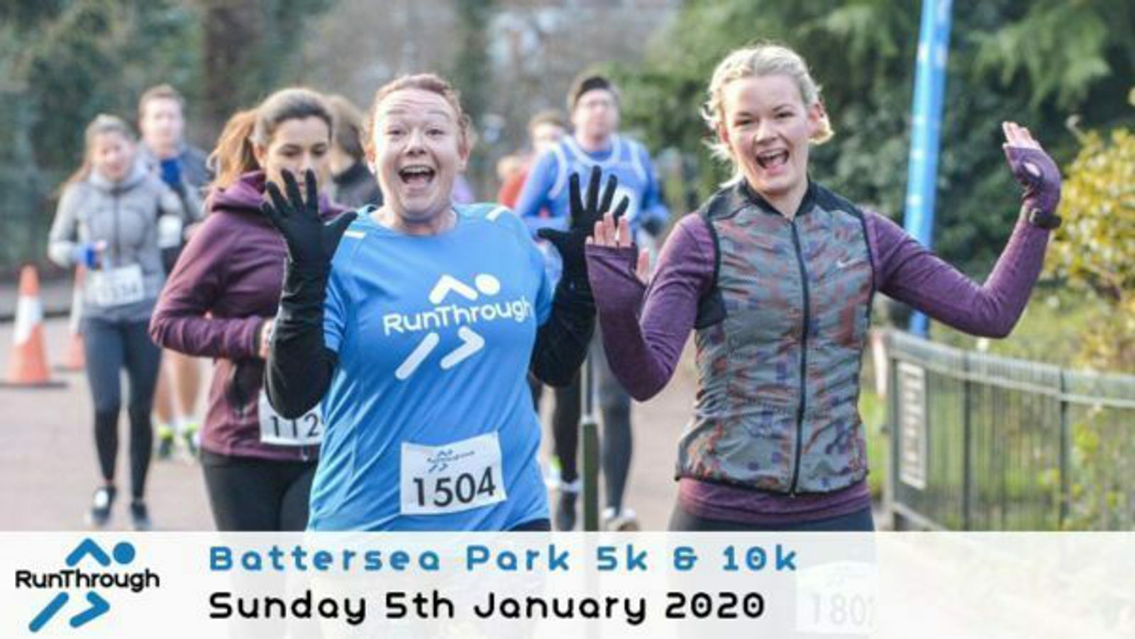 Two joyful participants, one in a blue RunThrough shirt and the other in a patterned jacket, smile and wave during the Battersea Park 5k & 10k run on Sunday, January 5th, 2020. Other runners and a blue event banner are visible in the background.