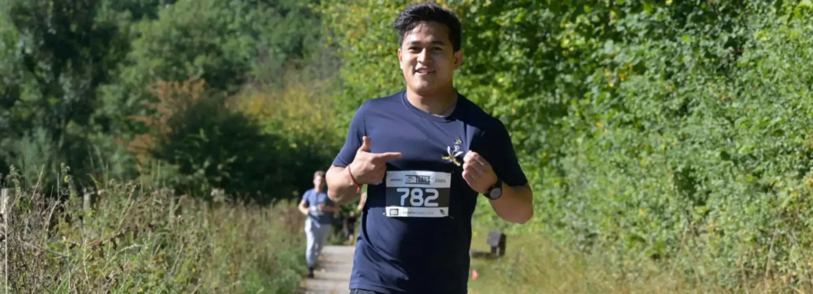 A man wearing a navy blue shirt, race bib 782, and a watch runs outdoors on a sunlit path lined with greenery, smiling and pointing at his bib. Other runners are visible in the background.