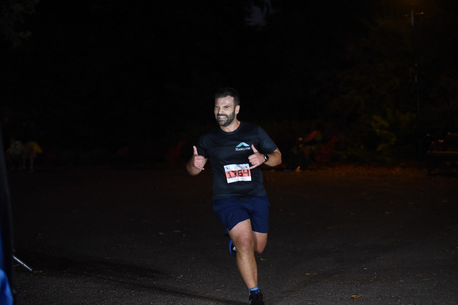 A man with a race bib number 1364 runs in a nighttime event, smiling and giving a thumbs-up gesture. He is dressed in dark athletic sportswear. The background is dimly lit with faint streetlights and trees visible.