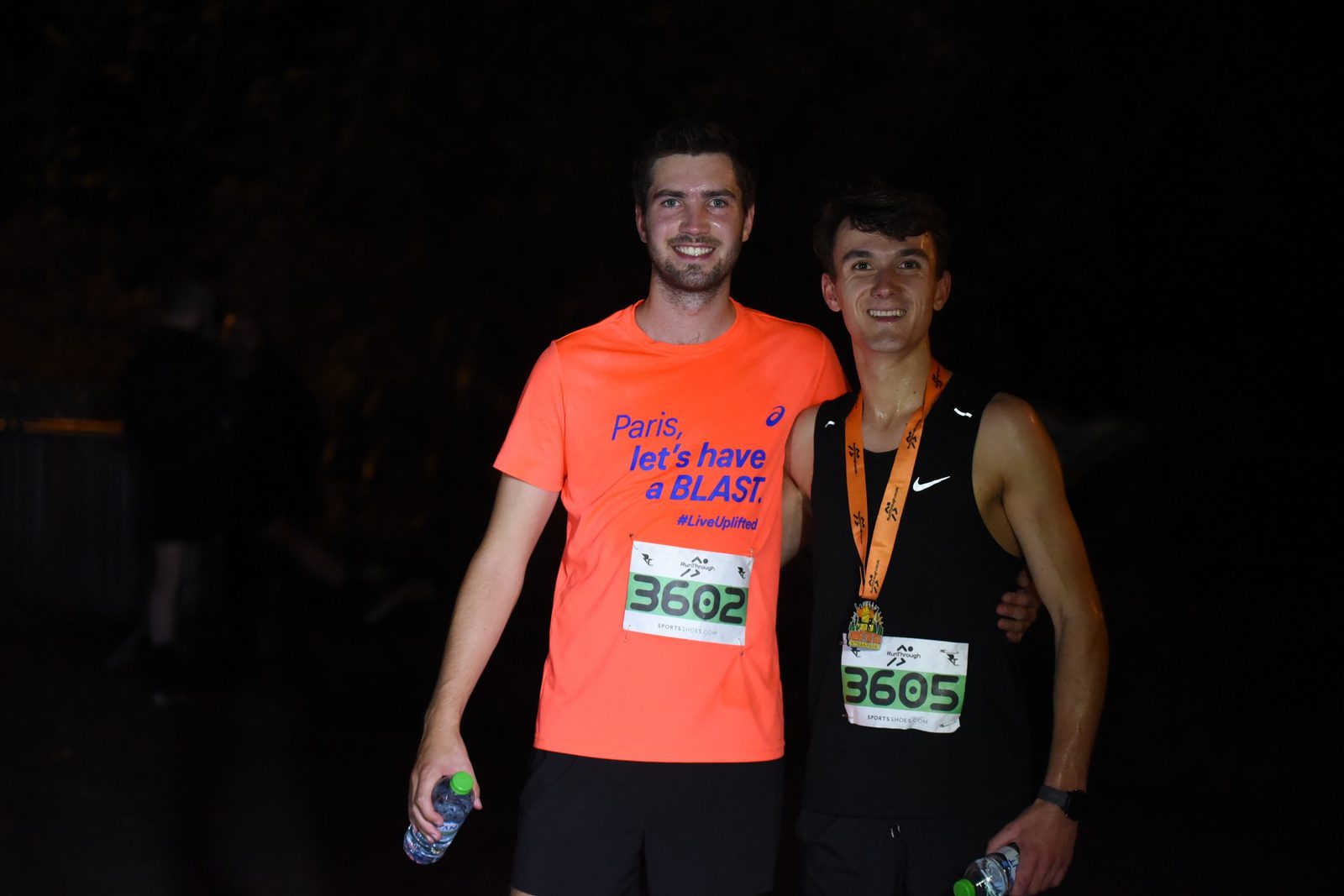 Two men posing together at a sports event at night. The man on the left is wearing an orange shirt and holding a water bottle, with bib number 3602. The man on the right is in a black tank top, holding a water bottle, with bib number 3605 and a medal around his neck.