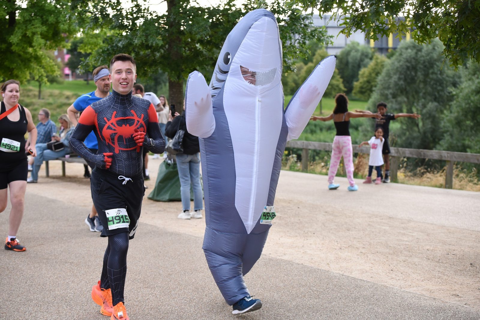 A person in a Spider-Man costume and another in an inflatable shark costume run side by side in a park during a fun run event. Other participants and spectators are visible in the background amidst greenery and walking paths.