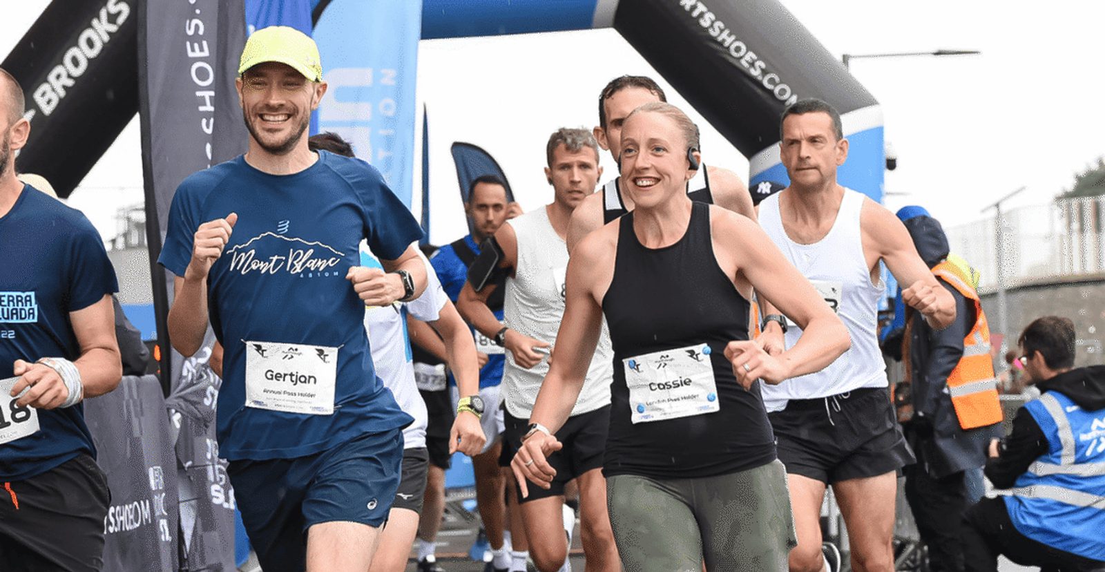 A group of runners, both men and women, wearing race bibs, smile as they start a race under an inflatable arch on a cloudy day. A few race officials and volunteers are visible in the background.