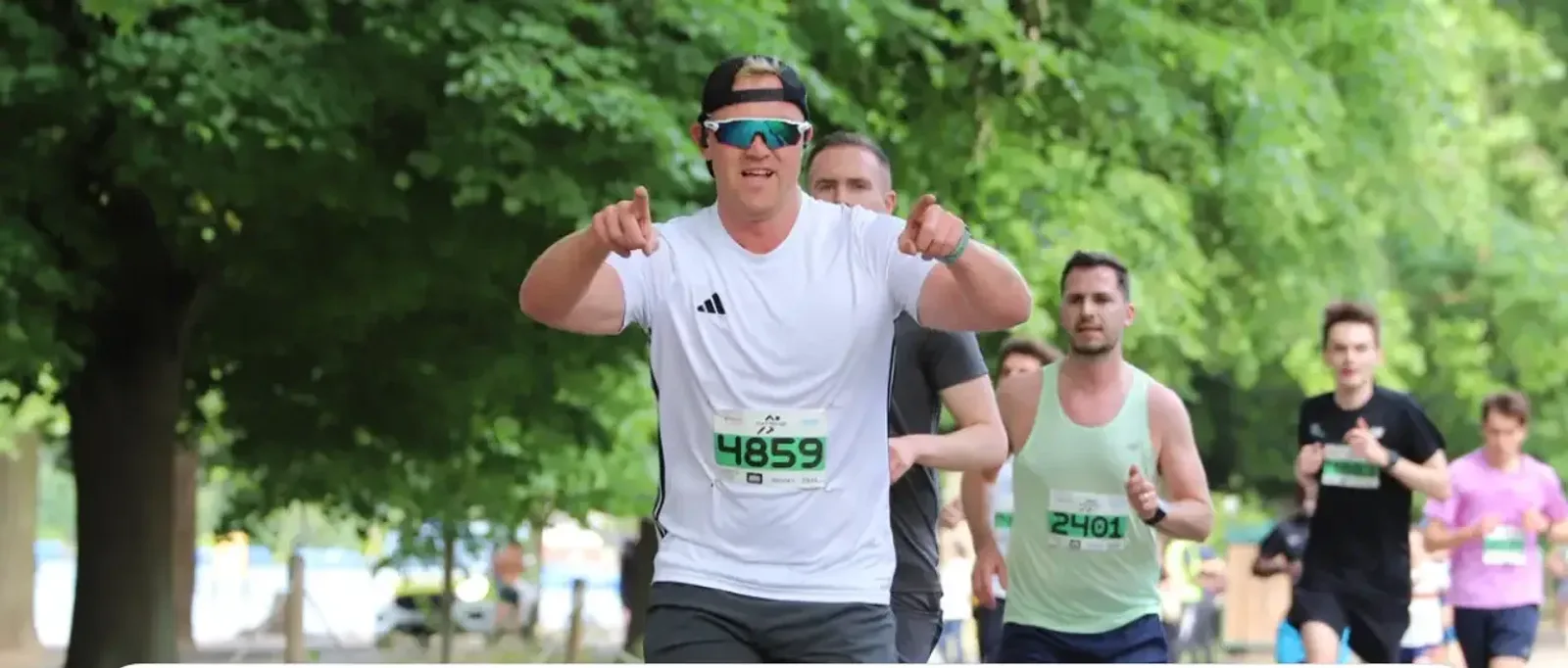 A man in a white shirt and sunglasses runs in a race, smiling and pointing at the camera. Several other runners, both male and female, follow behind him on a tree-lined path.