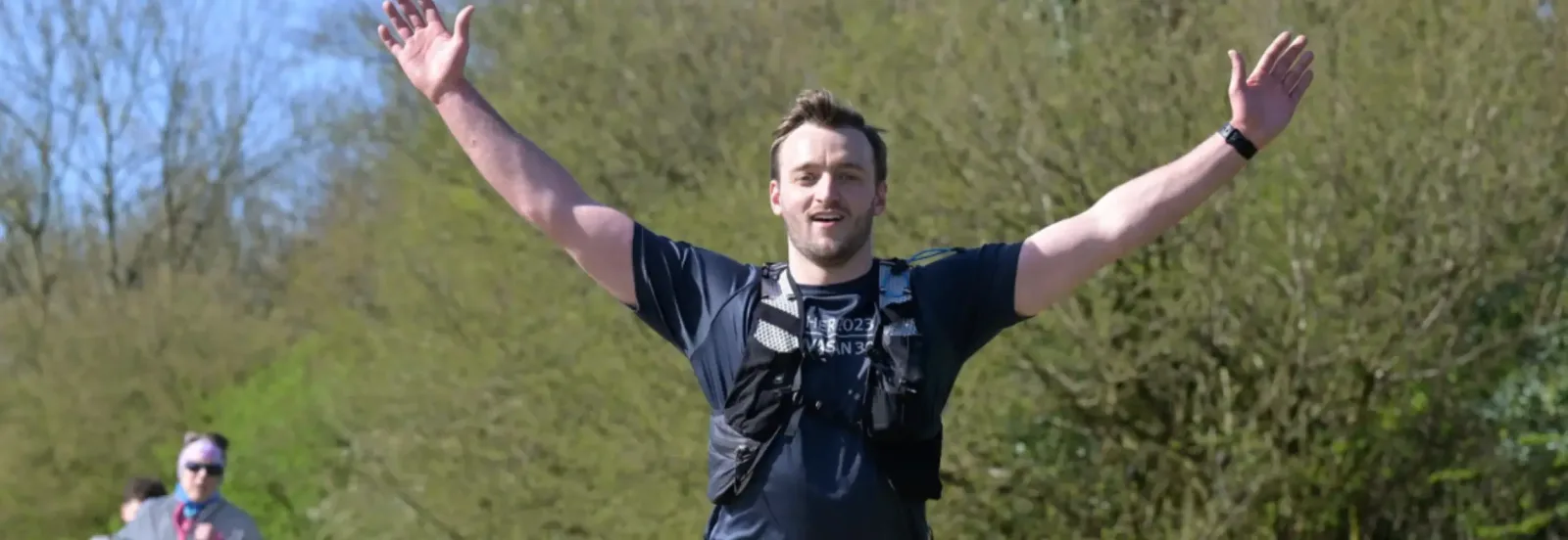 A man in a dark shirt and running vest raises both arms in celebration while running outdoors on a sunny day, with trees and blue sky in the background. Another runner is visible in the distance.
