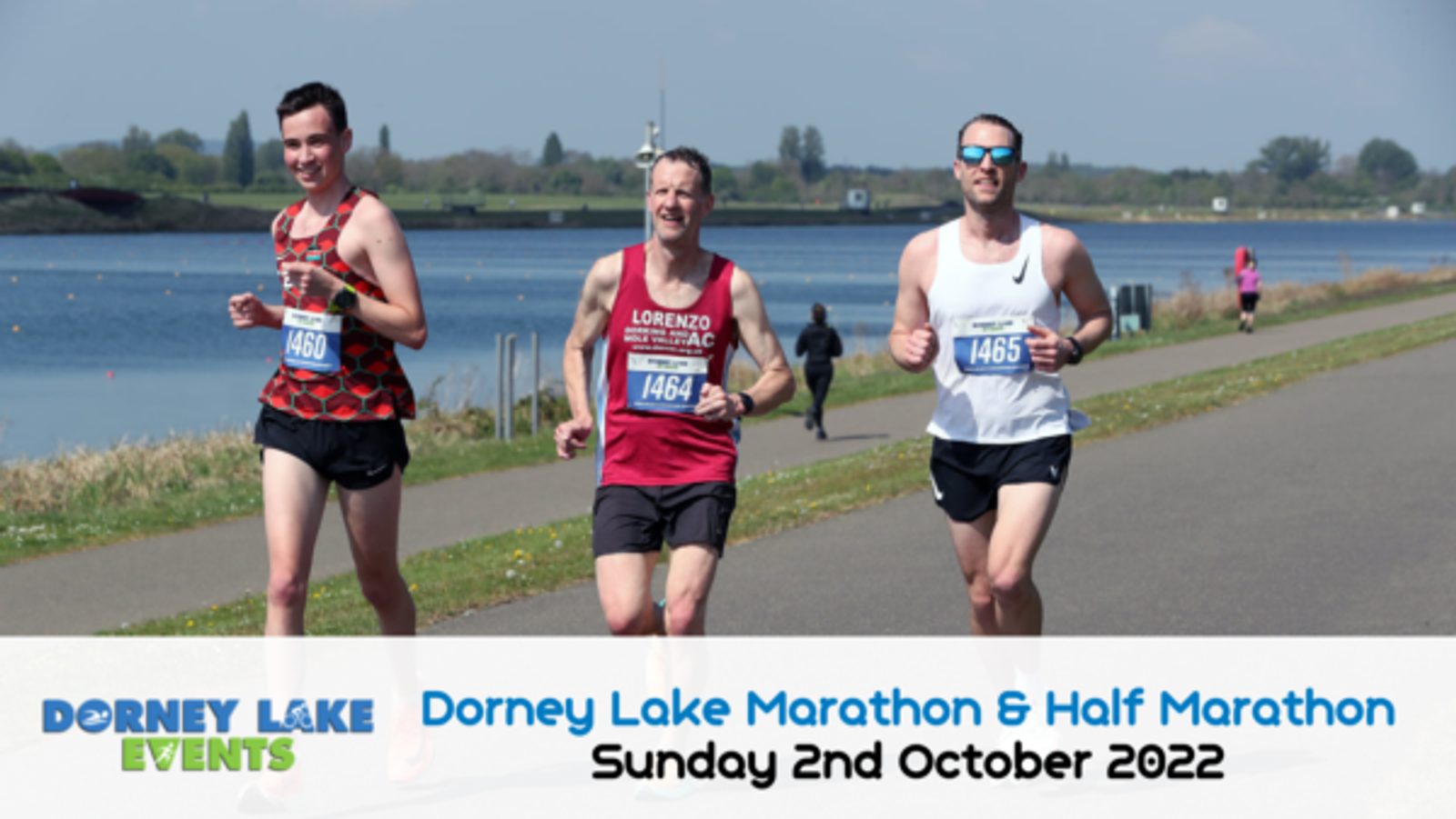 Three men are running along a paved path beside a lake during the Dorney Lake Marathon & Half Marathon on Sunday, 2nd October 2022. Two runners wear red shirts, and one wears a white shirt. Blue skies and green grass border the lake in the background.