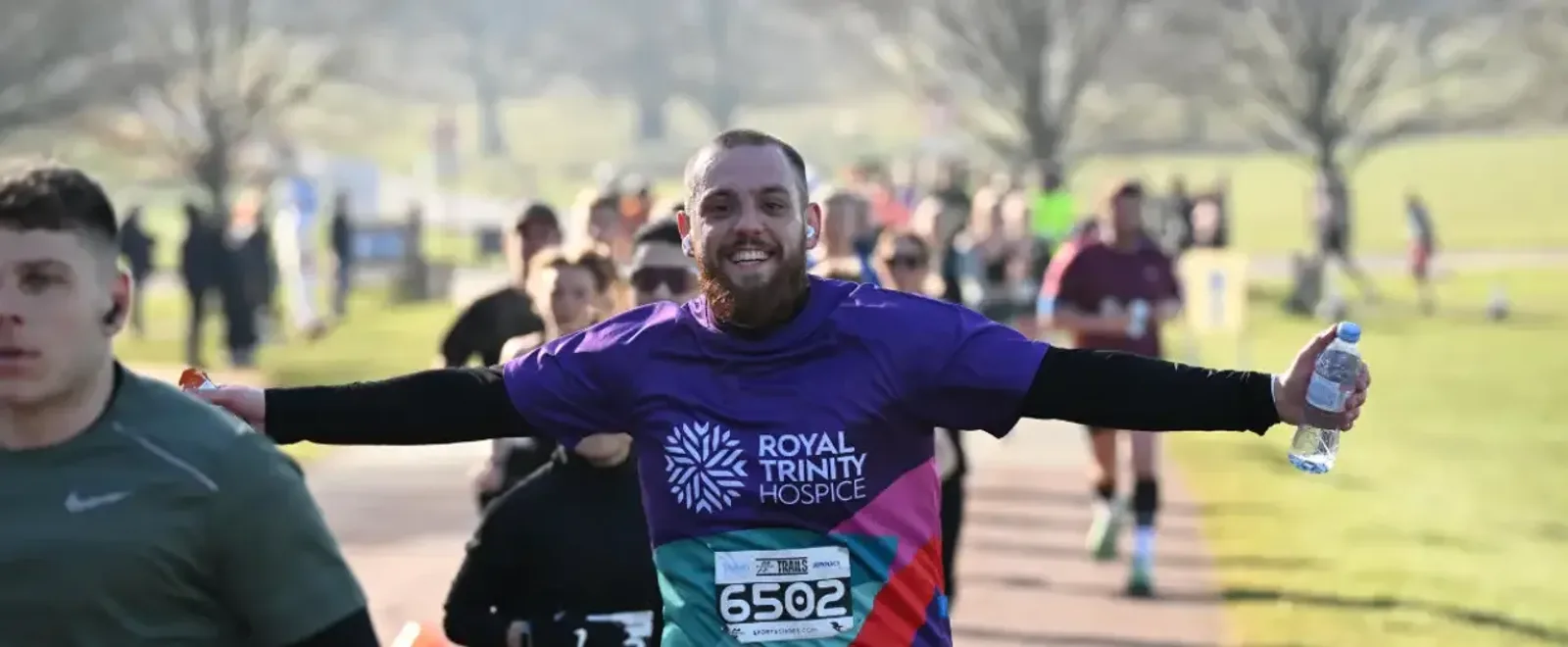 A smiling man in a purple "Royal Trinity Hospice" shirt runs with arms outstretched, holding a water bottle. Other runners are visible in the background on a sunlit park path.