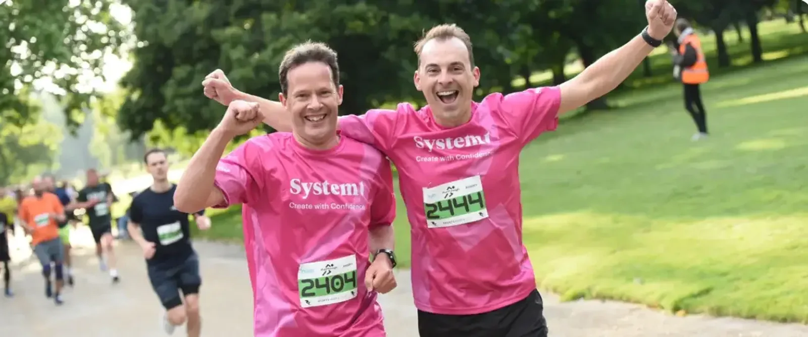 Two smiling men in pink shirts with race bibs run side by side in a park, arms raised in celebration. Other runners are visible in the background on a sunny day.