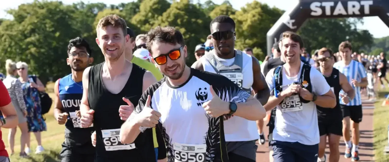 A group of smiling runners, several giving thumbs up, begin a race at the starting line on a sunny day, with trees and cheering spectators in the background.