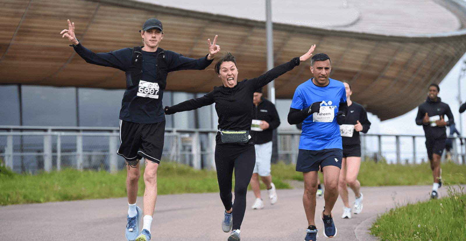 Three runners in athletic wear smile and gesture energetically as they run on a paved path during a race, with several other runners and a modern building visible in the background.