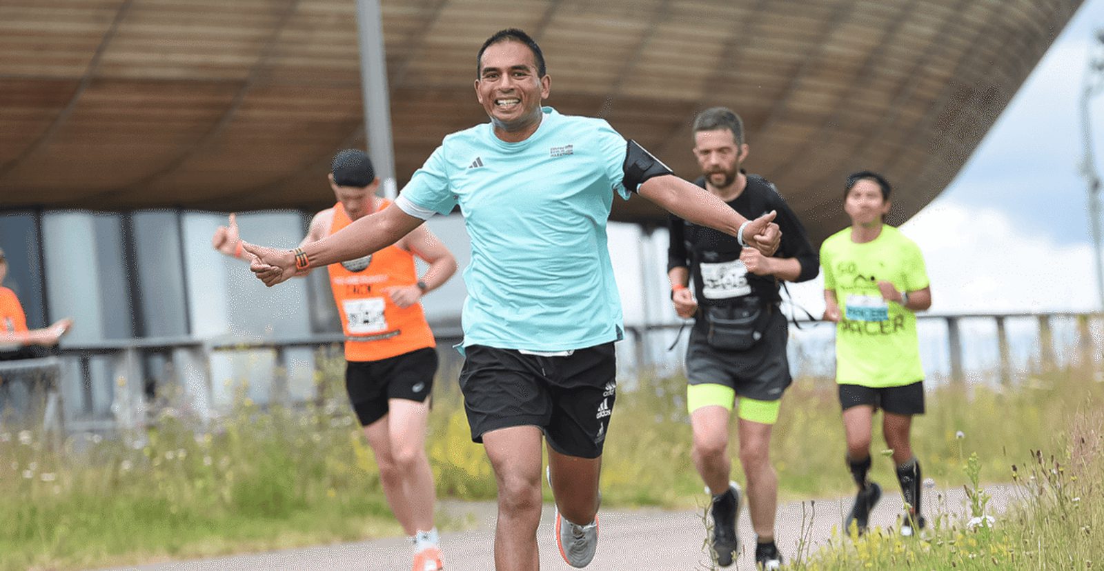 A smiling runner in a light blue shirt spreads his arms wide while running on a path, with other runners behind him. Tall grass and a modern building are visible in the background.