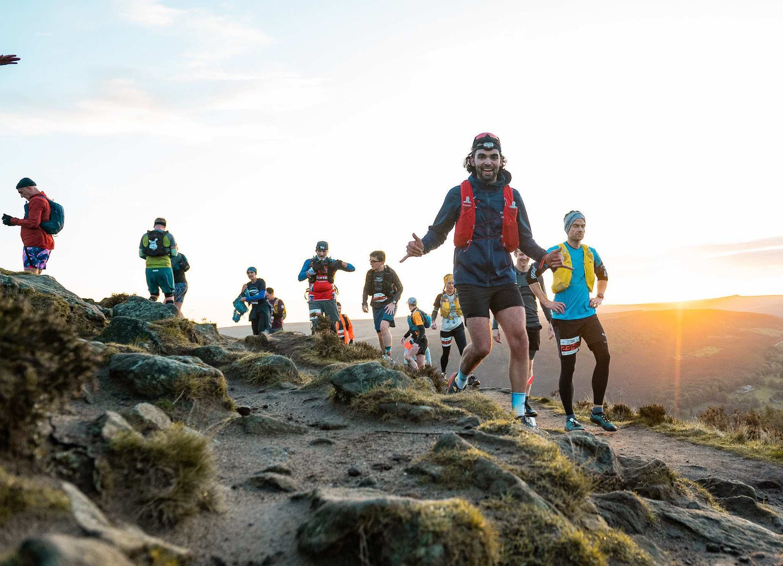 A group of trail runners, wearing race bibs and colorful gear, navigate a rocky path on a hillside during sunset. The setting sun casts a warm glow over the scenic landscape in the background.