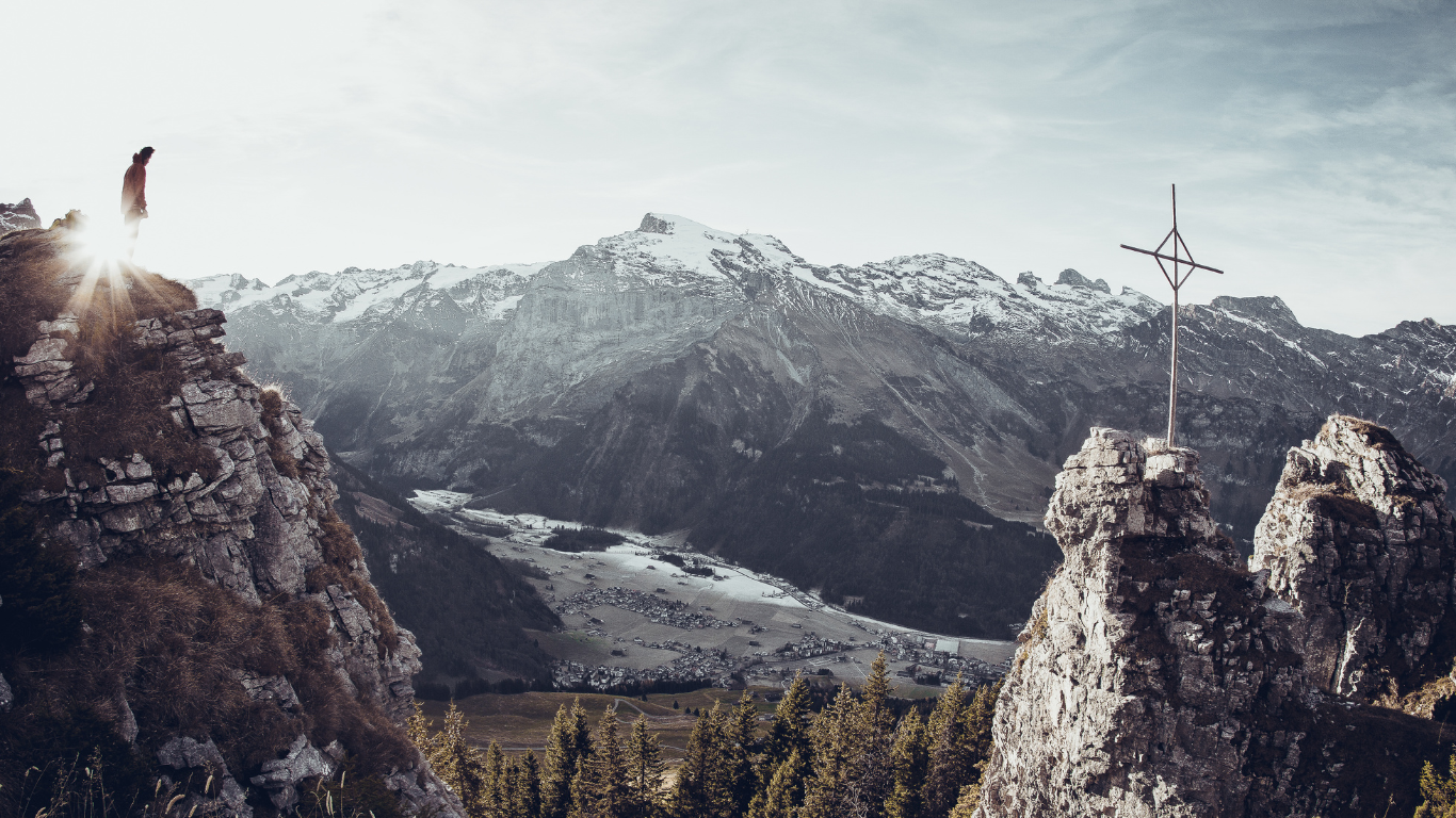 A person stands on a rocky cliff with the sun shining behind them, overlooking a valley and snow-capped mountain range. A cross is visible on a nearby peak, and a small village is nestled in the valley below.