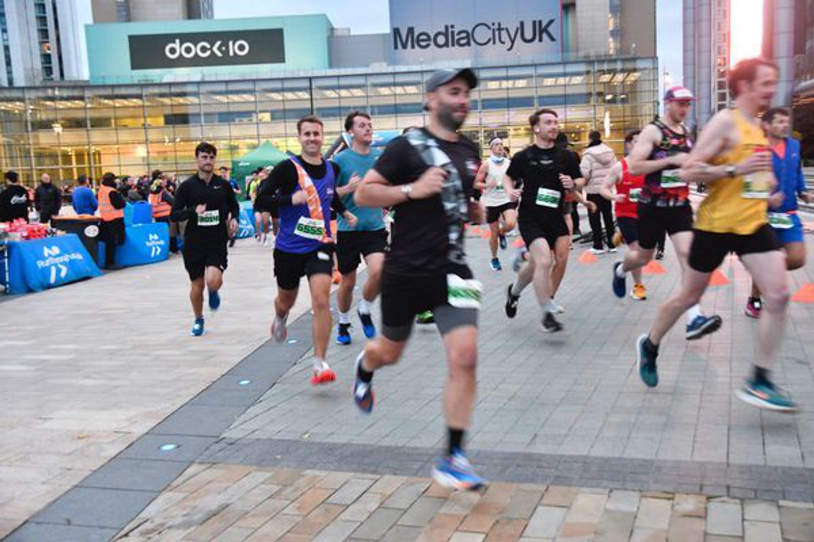 Runners in a race pass by a modern building with "MediaCityUK" and "dock10" signage. The scene is dynamic, capturing energy and motion as the athletes stride forward on a paved path.