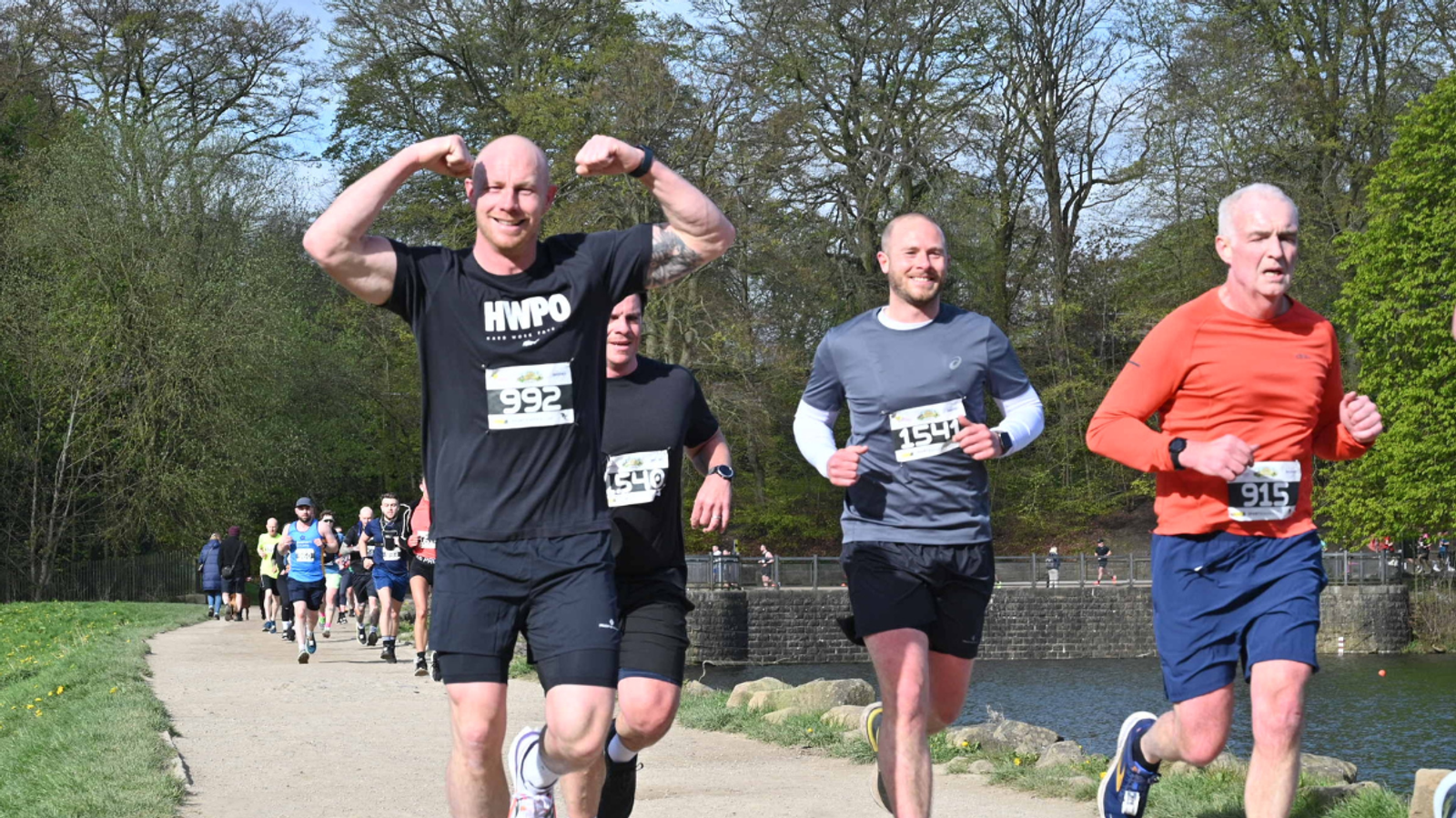 A group of runners participating in a race along a scenic path beside a river. One man in a black shirt is in the foreground, flexing his muscles and smiling. Others follow behind, with trees and a sunny sky in the background.