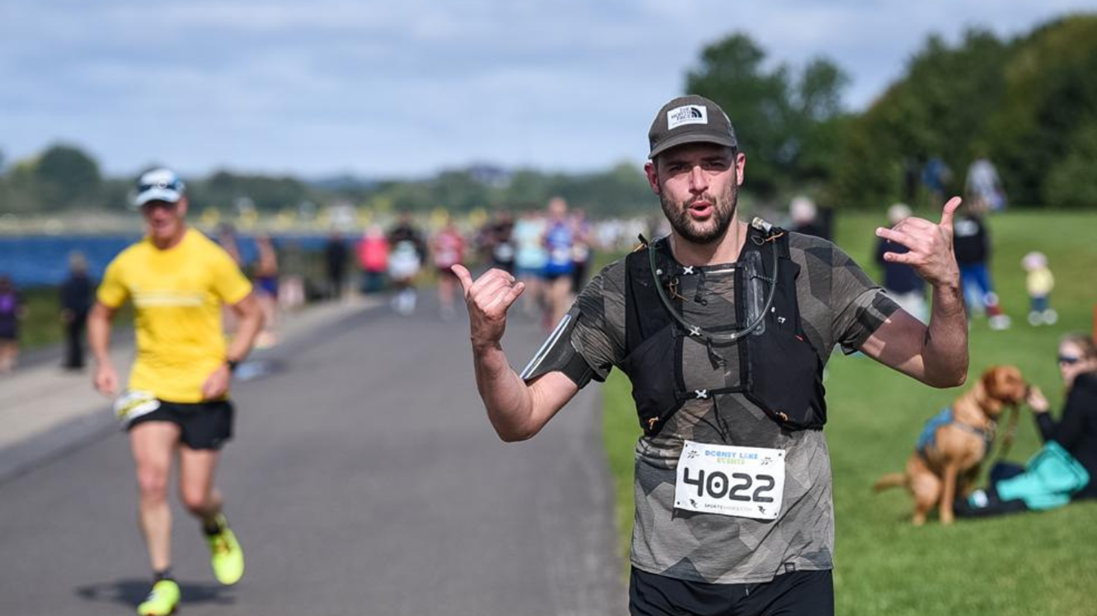 A runner wearing a gray shirt, cap, and bib number 4022 gestures enthusiastically while running on a park path. Other runners and spectators are visible in the background, with a dog and its owner on the grass.